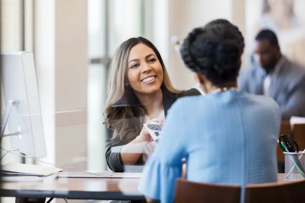 a woman in an office handing another woman a pamphlet. 